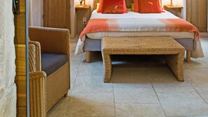 Un dormitorio con una cama, dos lámparas de noche, una silla de mimbre, un banco de madera y un cuadro de un paisaje urbano en la pared.