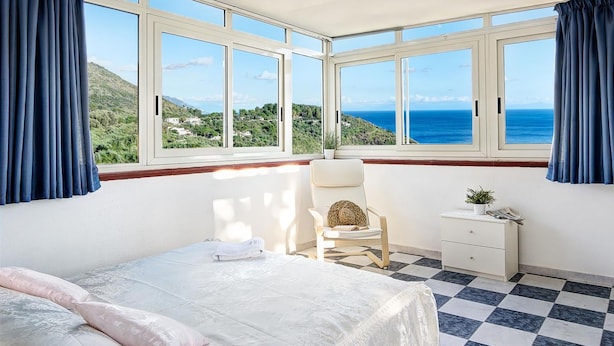 A bedroom with a large bay window, a bed, a chair, a nightstand, and a view of the sea and hills. - Casa Maradona (Massa Lubrense)