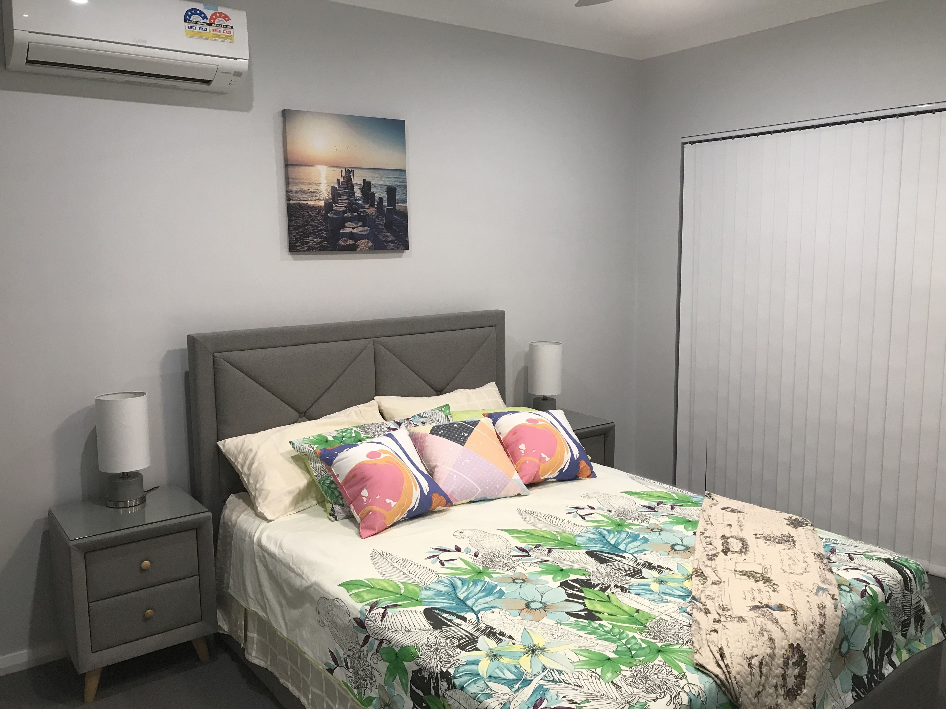 A bedroom with a bed, nightstands, a wall-mounted air conditioner, and a picture of a sunset over a pier.