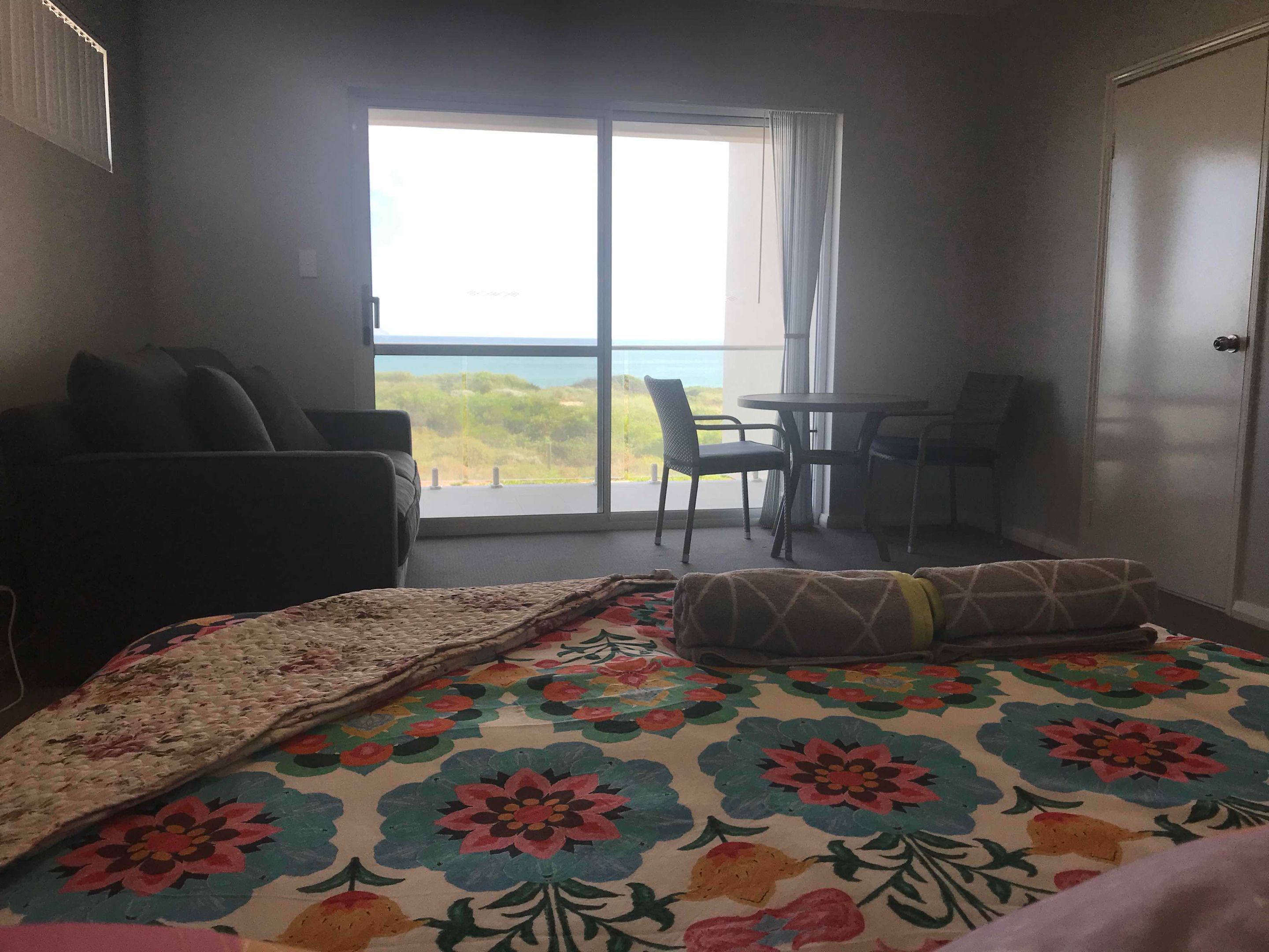 A hotel room with a bed, a small table with chairs, and a balcony with a view of the sea.