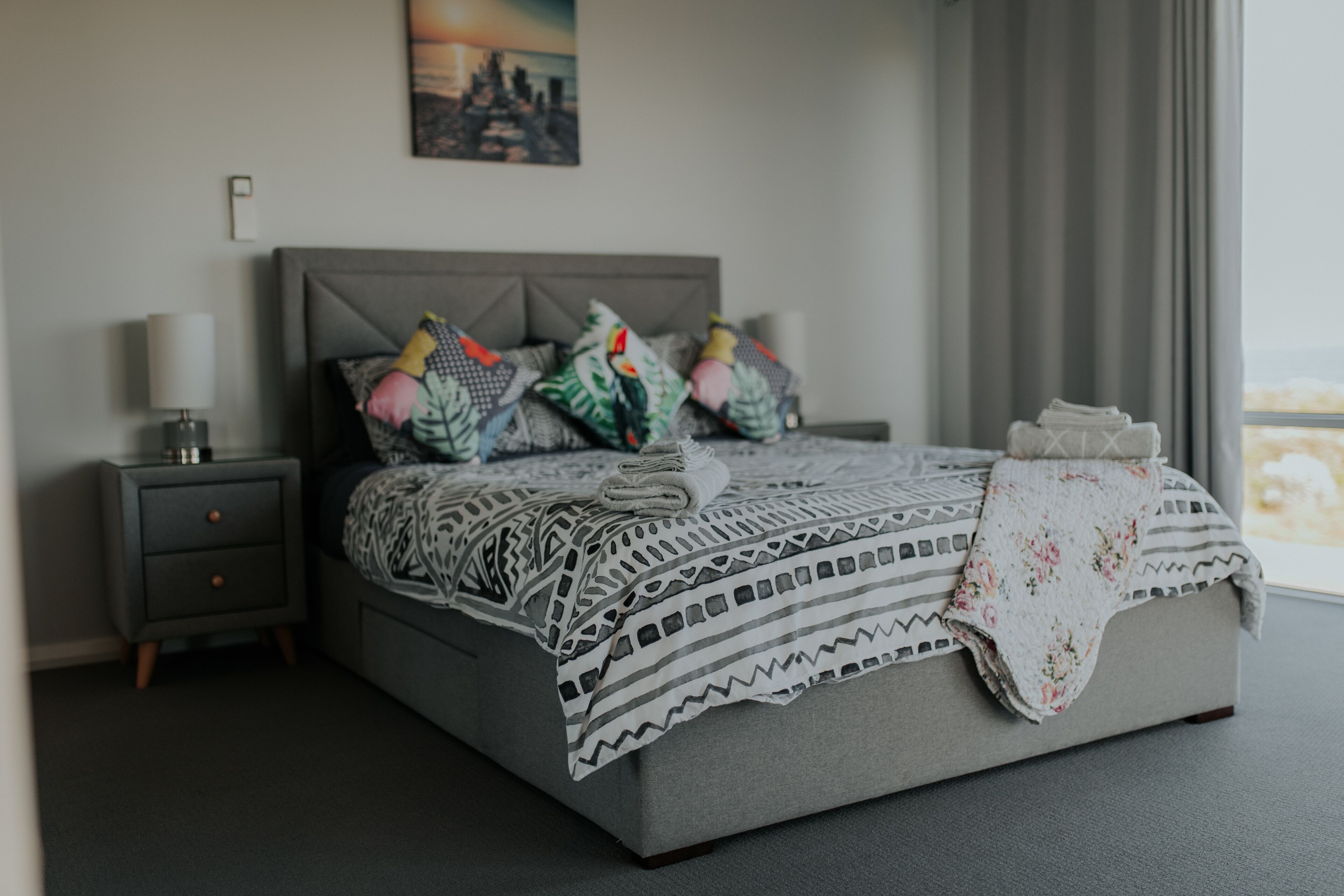 A neatly made bed with patterned bedding and pillows, a bedside table with a lamp, and a window with curtains.