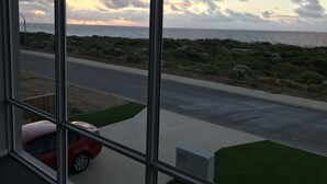 Vista a través de una ventana mostrando una carretera costera, un coche rojo estacionado al lado, un área con césped y el mar al fondo.