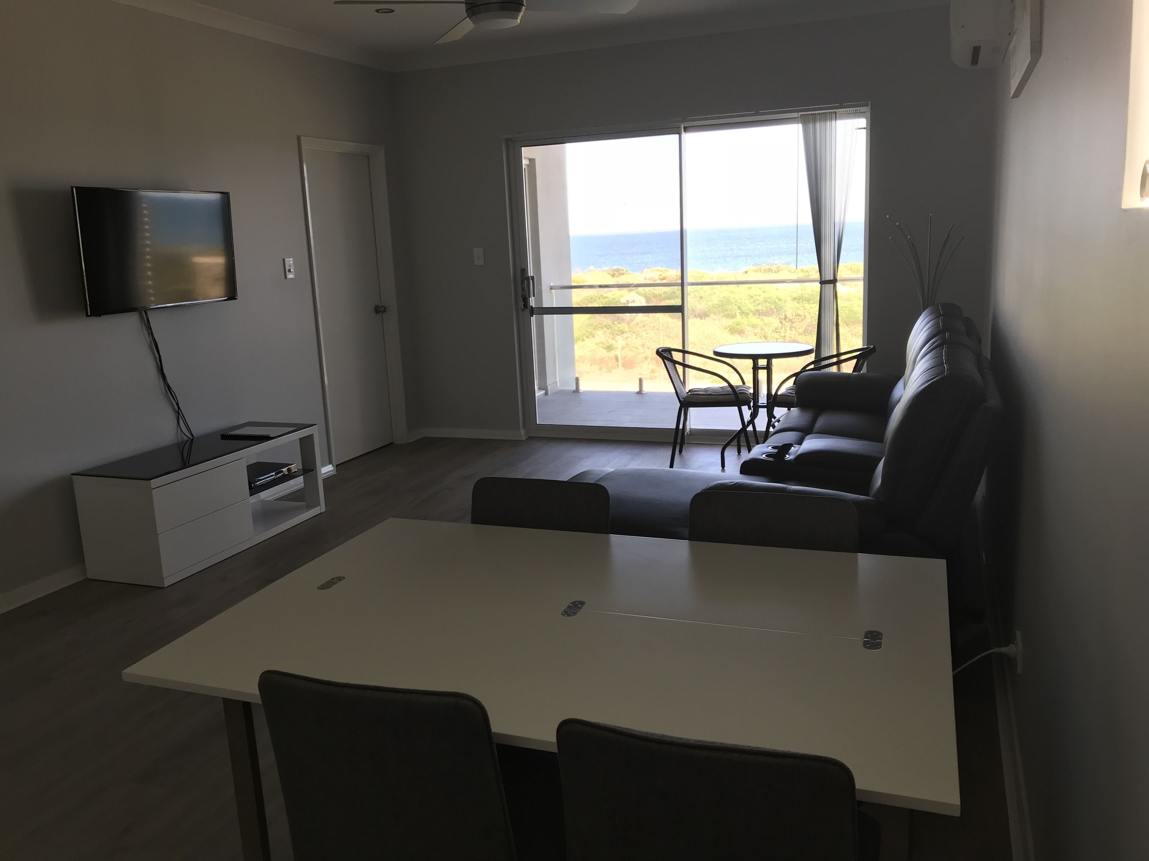 A modern living room with a flat-screen TV, a sofa, a dining table, and a view of the beach.