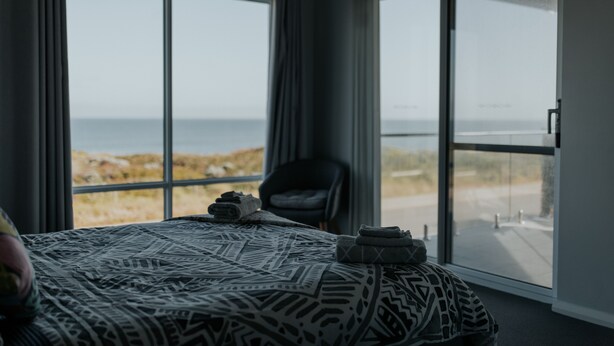 Un dormitorio con cama, una silla y ventanas amplias con vista a la playa.