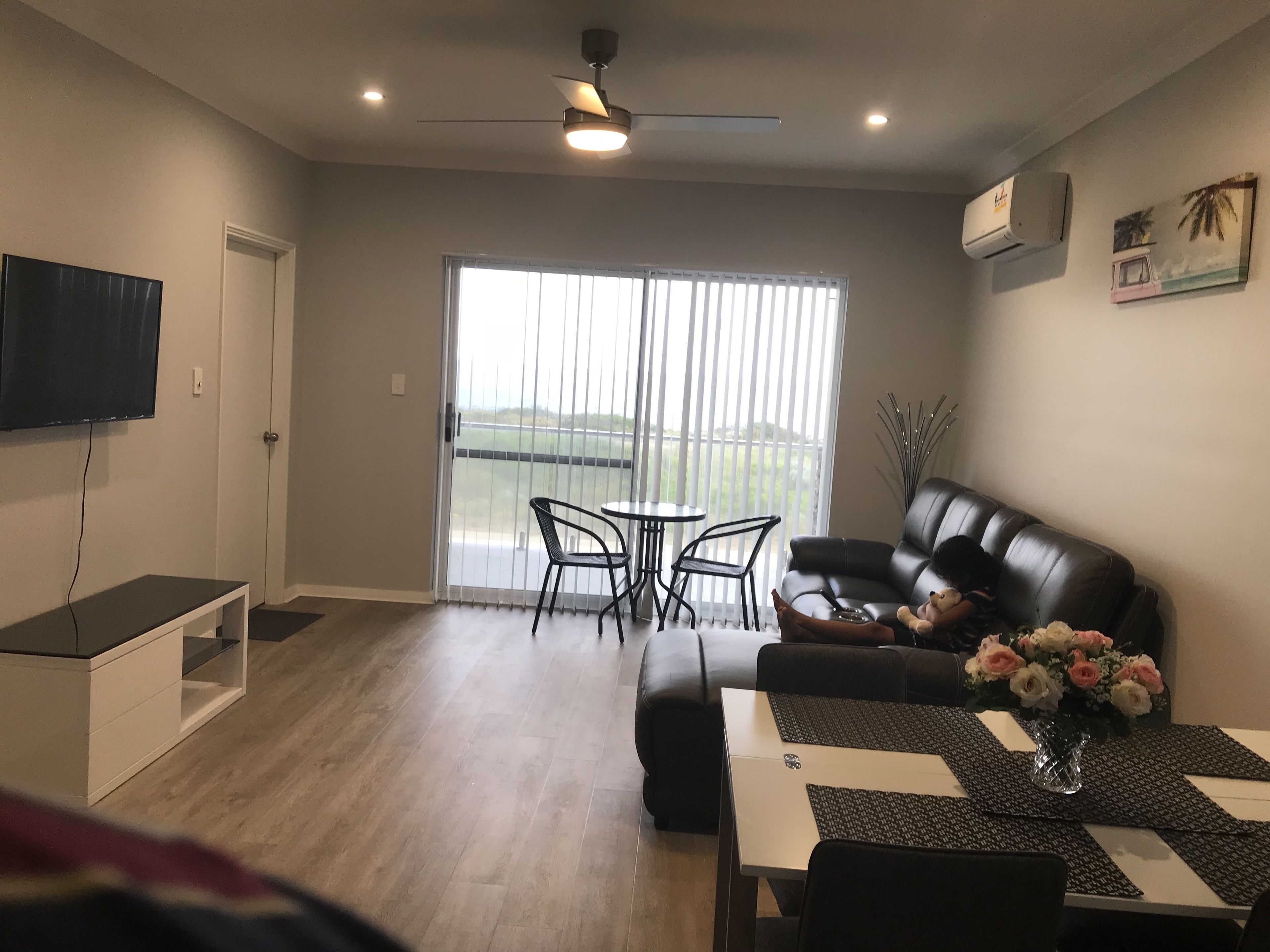 Uma sala de estar com um sofá de couro preto, uma TV na parede, uma mesa de jantar com jogos americanos e uma varanda com vista.