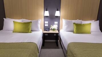 Habitación de hotel con dos camas, cada una con ropa de cama blanca y una almohada verde. Entre las camas hay una mesita con una lámpara y una pequeña planta.