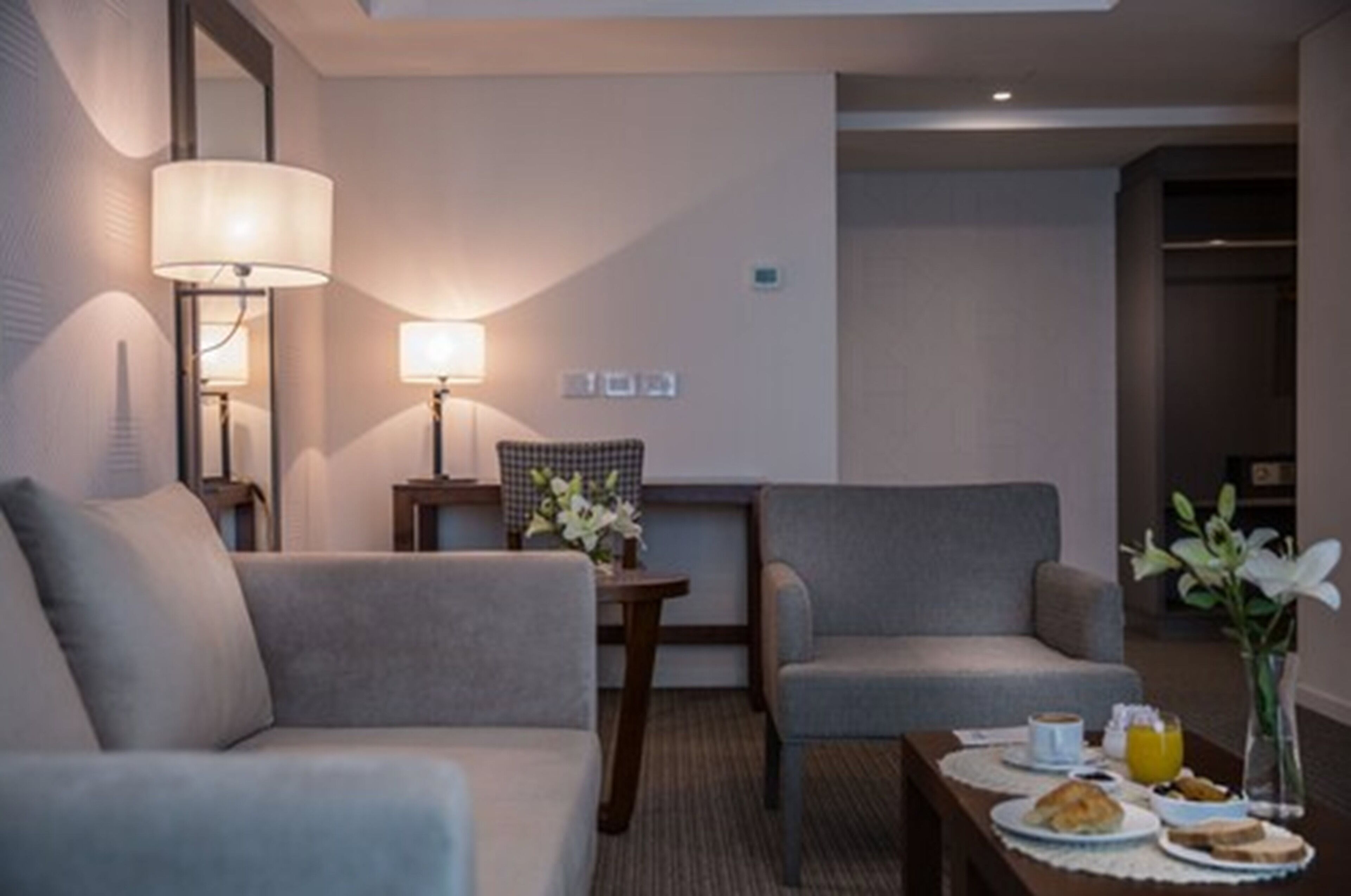 A hotel room with a sofa, two armchairs, a small table with breakfast items, and a lamp.