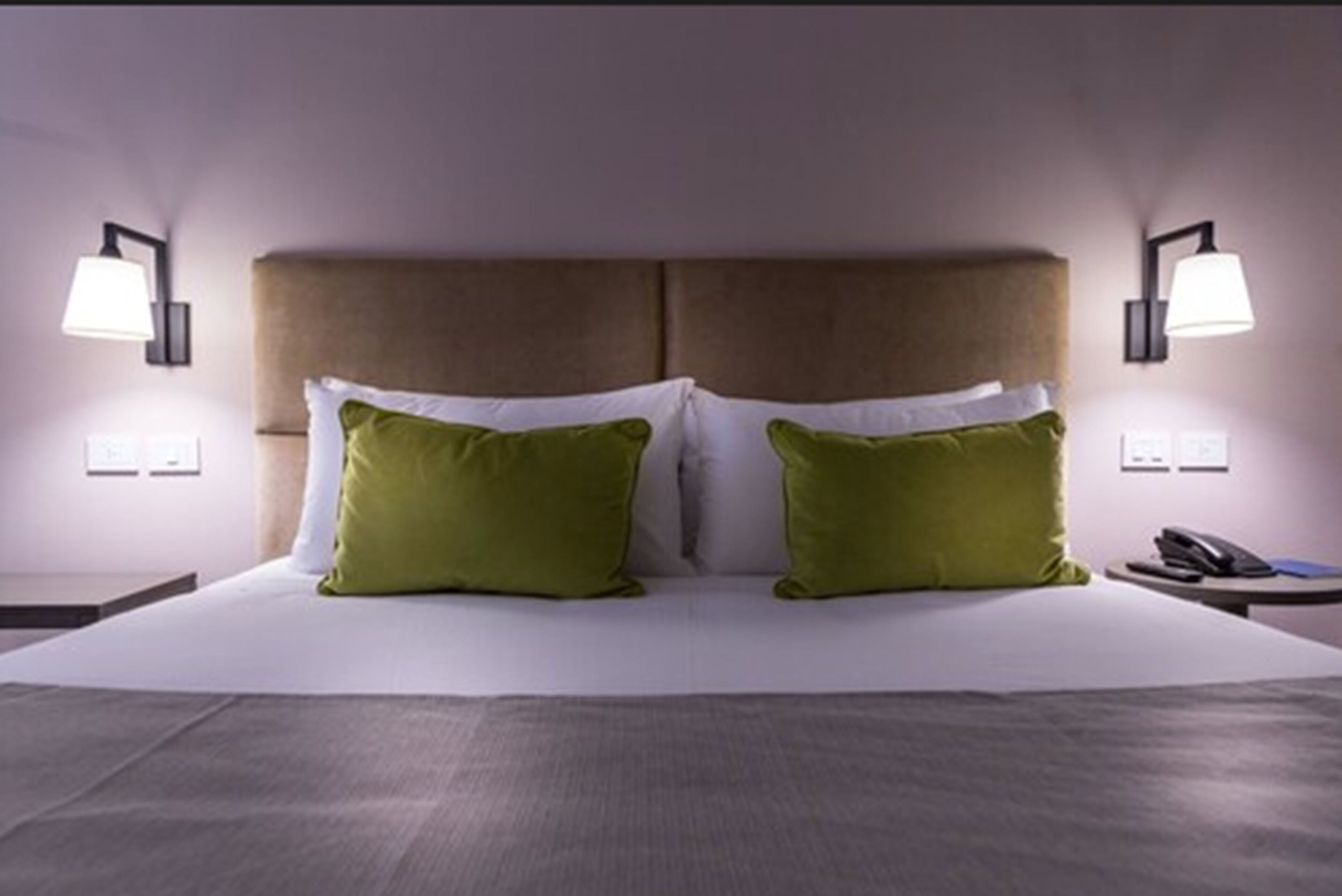A hotel room with a large bed, two green pillows, a headboard, bedside lamps, and a phone on the nightstand.
