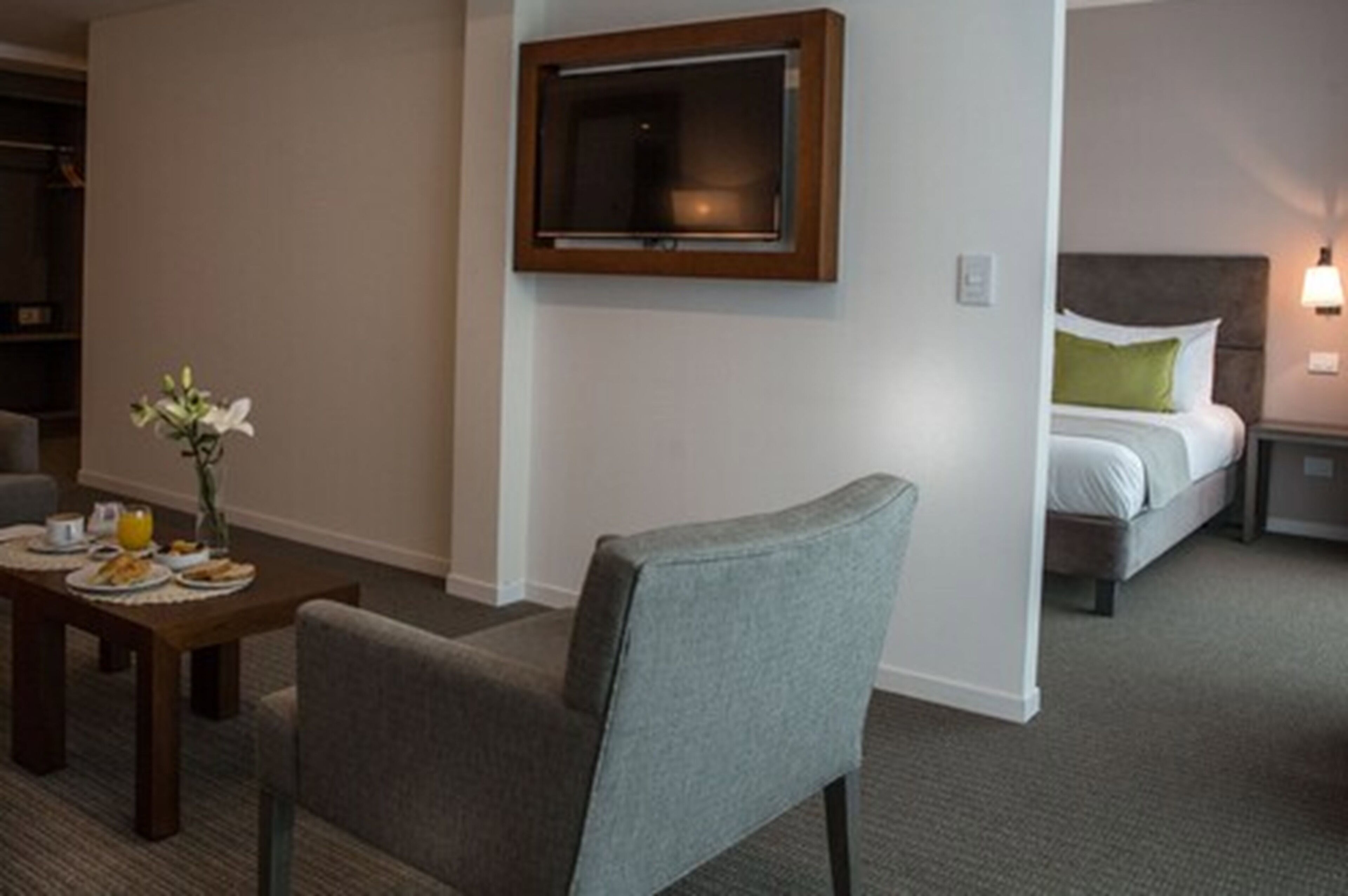 A hotel room with a bed, a bedside table with a lamp, a chair, and a TV mounted on the wall.