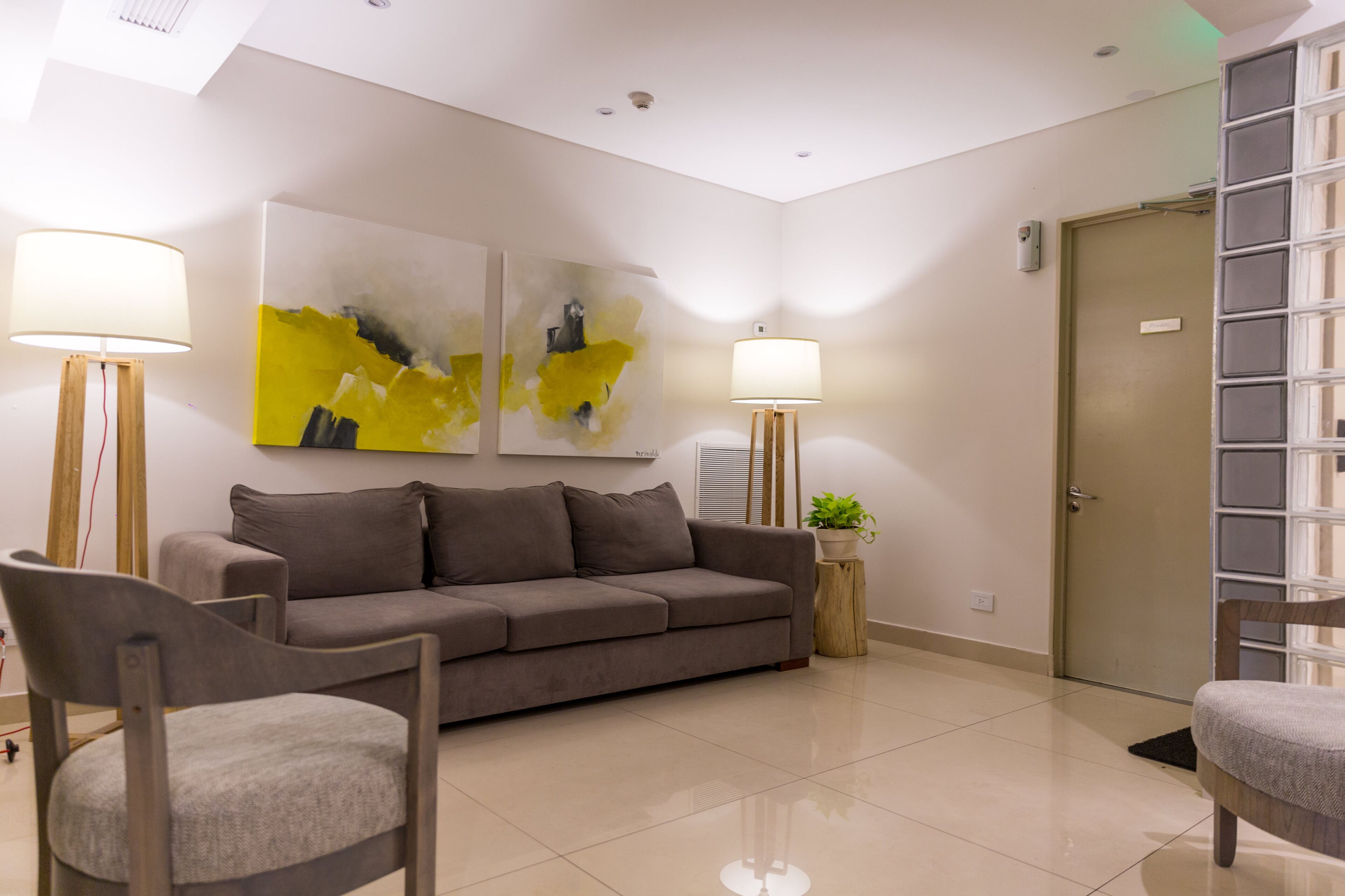 Una sala moderna con un sofá, dos sillones, una mesita de centro y dos lámparas de pie. Se observa una puerta y una ventana en el lado derecho.