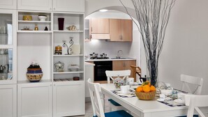 A modern kitchen with white cabinetry, a dining area with a white table set for two, and a decorative vase with branches. - Matisse Cantone (Massa Lubrense)