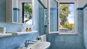 A bathroom with a white sink, a mirror, and a window with a view of trees and a fence. - Miomar (Massa Lubrense)
