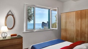 A bedroom with a bed, a wooden wardrobe, a nightstand with a lamp, and a window with a view of the sea. - Miomar (Massa Lubrense)