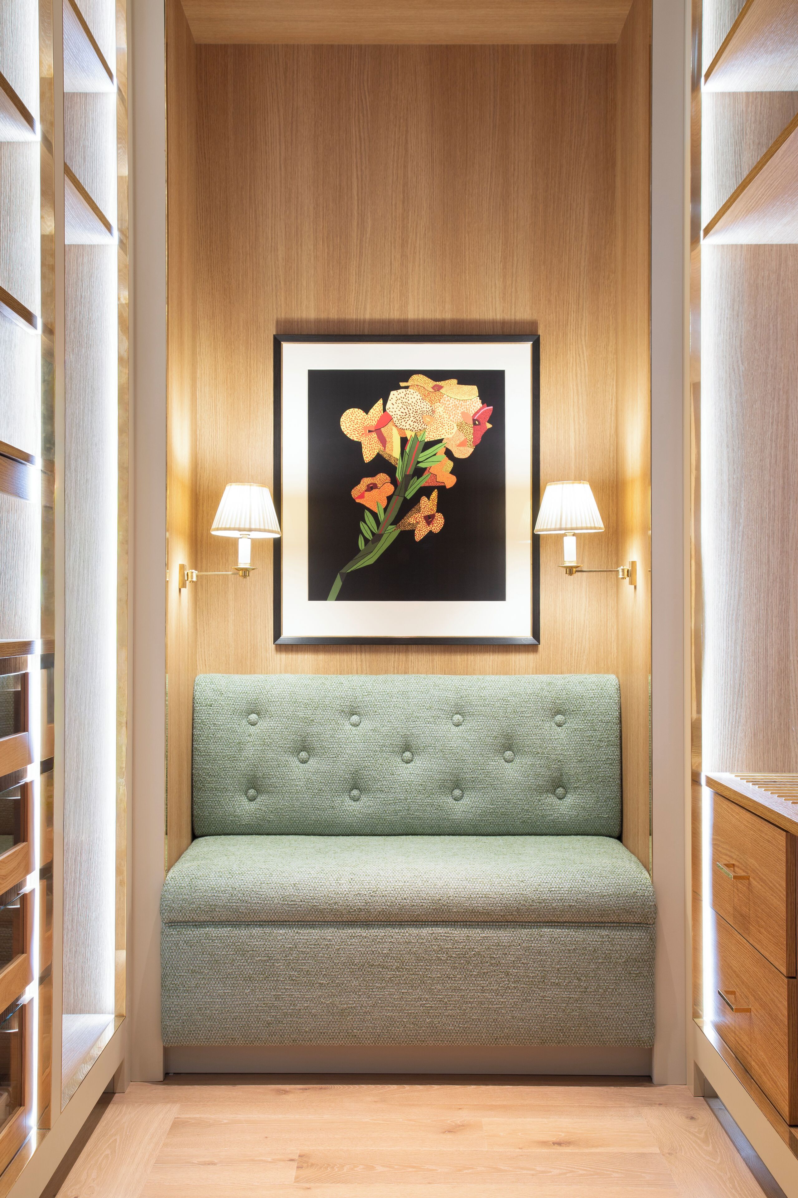 Un couloir avec un banc recouvert d’un tissu vert, des œuvres d’art florales encadrées au-dessus, et des lampes fixées aux murs de part et d’autre.