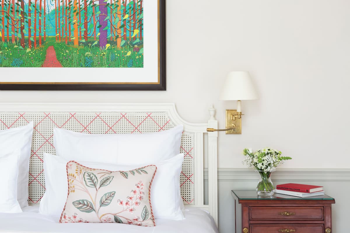 A neatly made bed with white and floral pillows, a framed colorful painting above it, a bedside lamp, a small wooden nightstand with a vase of flowers and a book, and a patterned headboard.