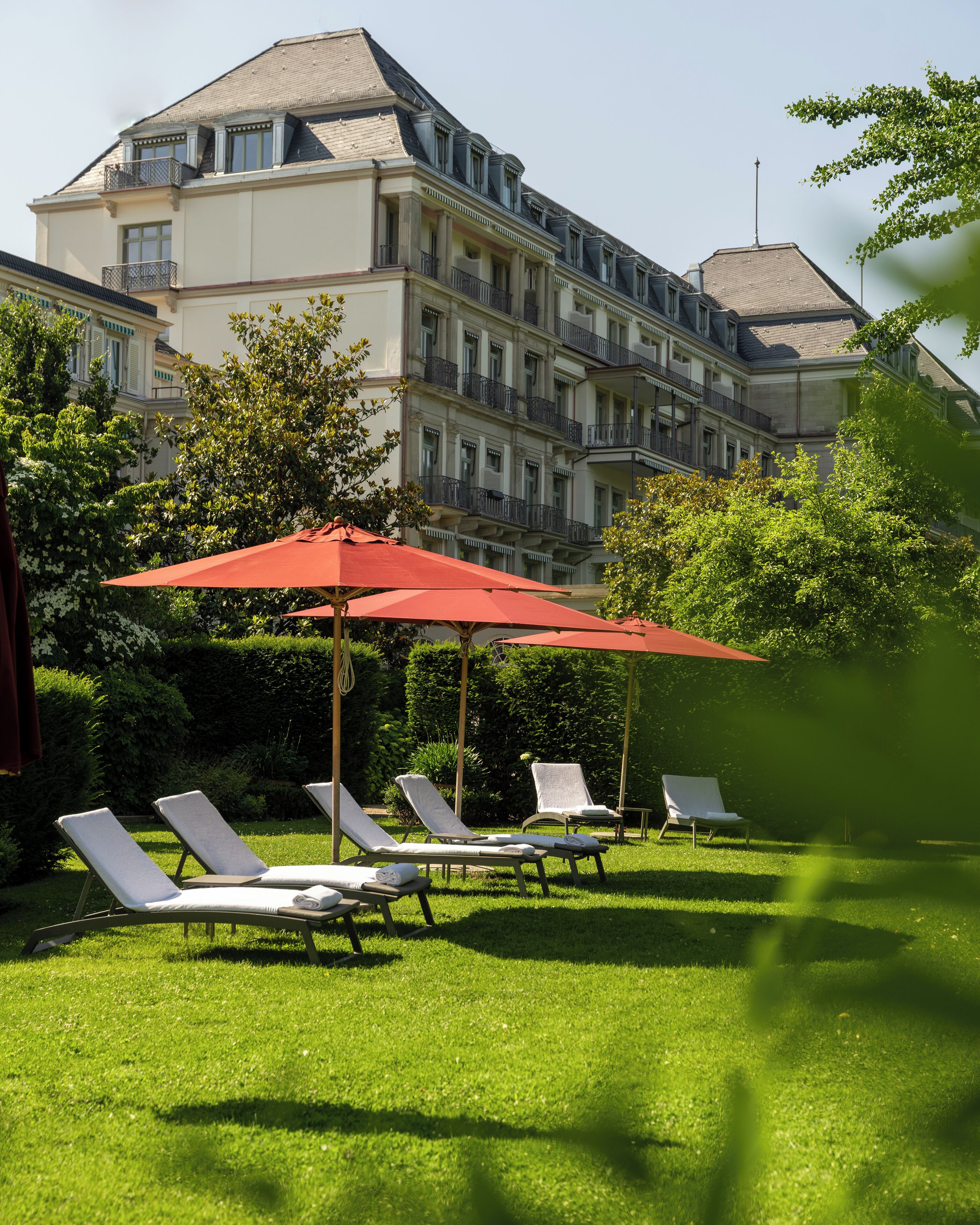 Ein Hotelinnenhof mit Liegestühlen und roten Sonnenschirmen, umgeben von üppigem Grün und einem mehrstöckigen Gebäude mit Balkonen.