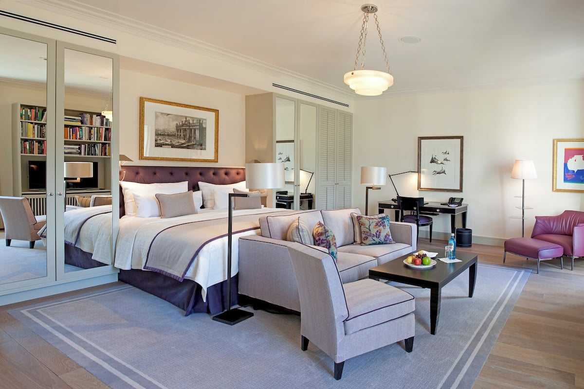 A modern hotel room with a large bed, a sofa, a desk, and a chair. There is a bookshelf and a mirror.