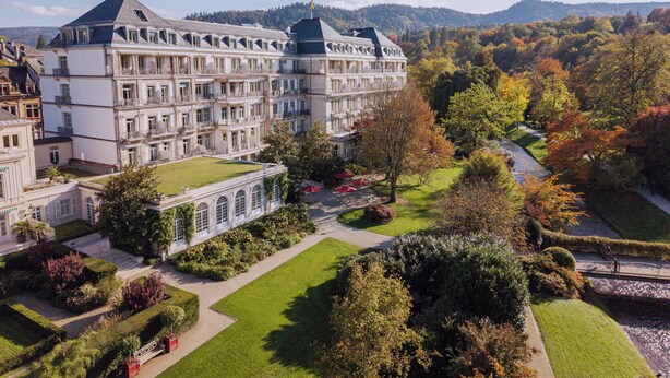 Un hotel grande de varios pisos con una fachada simétrica, extensos jardines y vistas a las colinas boscosas circundantes.