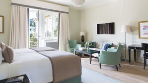 Habitación de hotel con una cama grande, zona de estar con una mesa y sillas, televisor montado en la pared y balcón con cortinas.