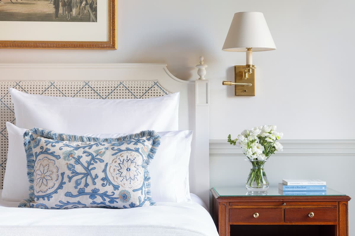 A neatly made bed with a patterned pillow, a bedside table with a vase of flowers, and a wall-mounted lamp.