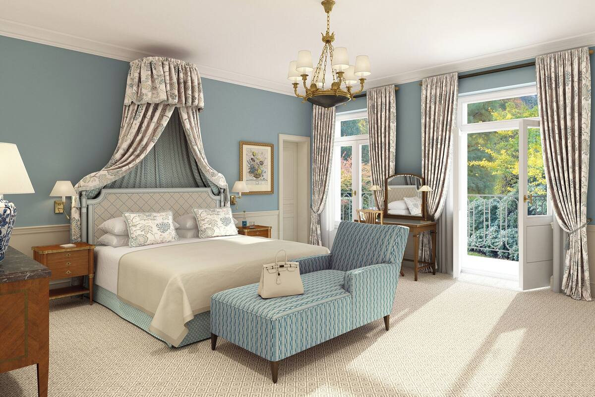 A bedroom with a canopy bed, a sitting area with a chair, a bedside table, a lamp, and a view of greenery outside.