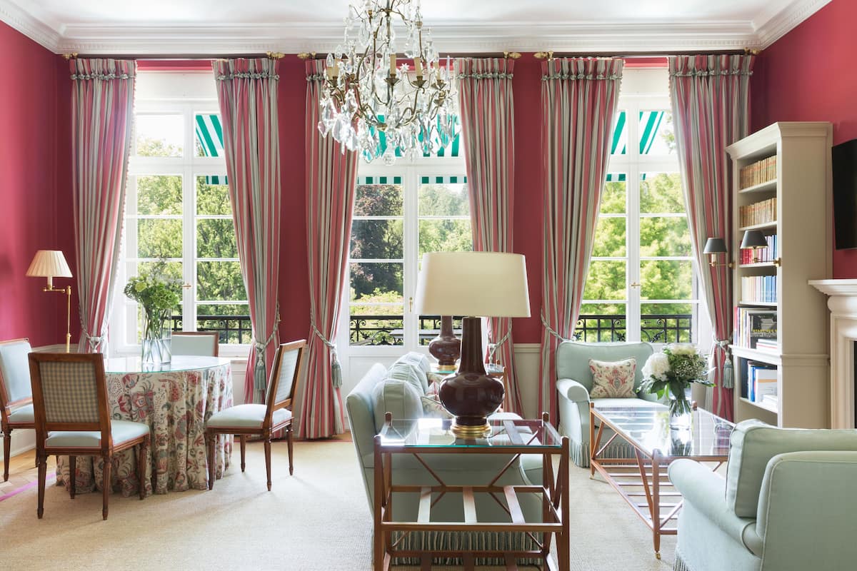 A room with a round table, chairs, a sofa, a bookshelf, and a large window with curtains.