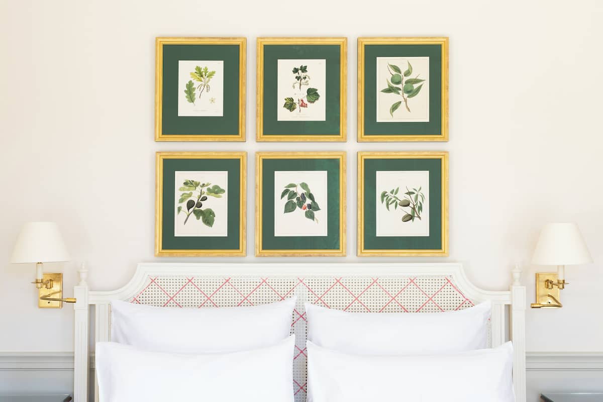 A neatly arranged bedroom with a bed, two wall-mounted lamps, and six framed botanical prints on the wall.