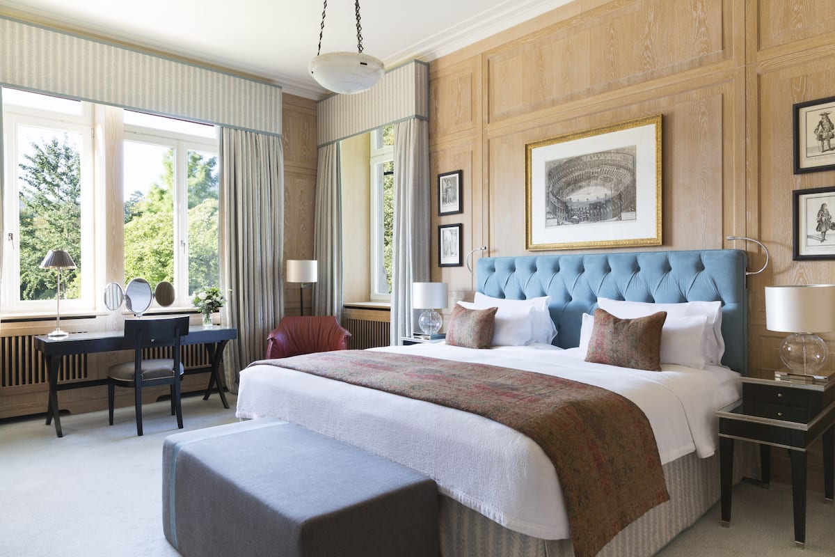 A well-appointed hotel room with a large bed, a desk with a chair, a window with curtains, and framed artwork on the walls.