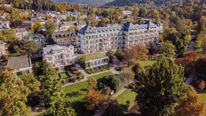 Un edifici gran i històric amb diverses plantes i una façana simètrica, envoltat de vegetació exuberant i un paisatge amb turons.