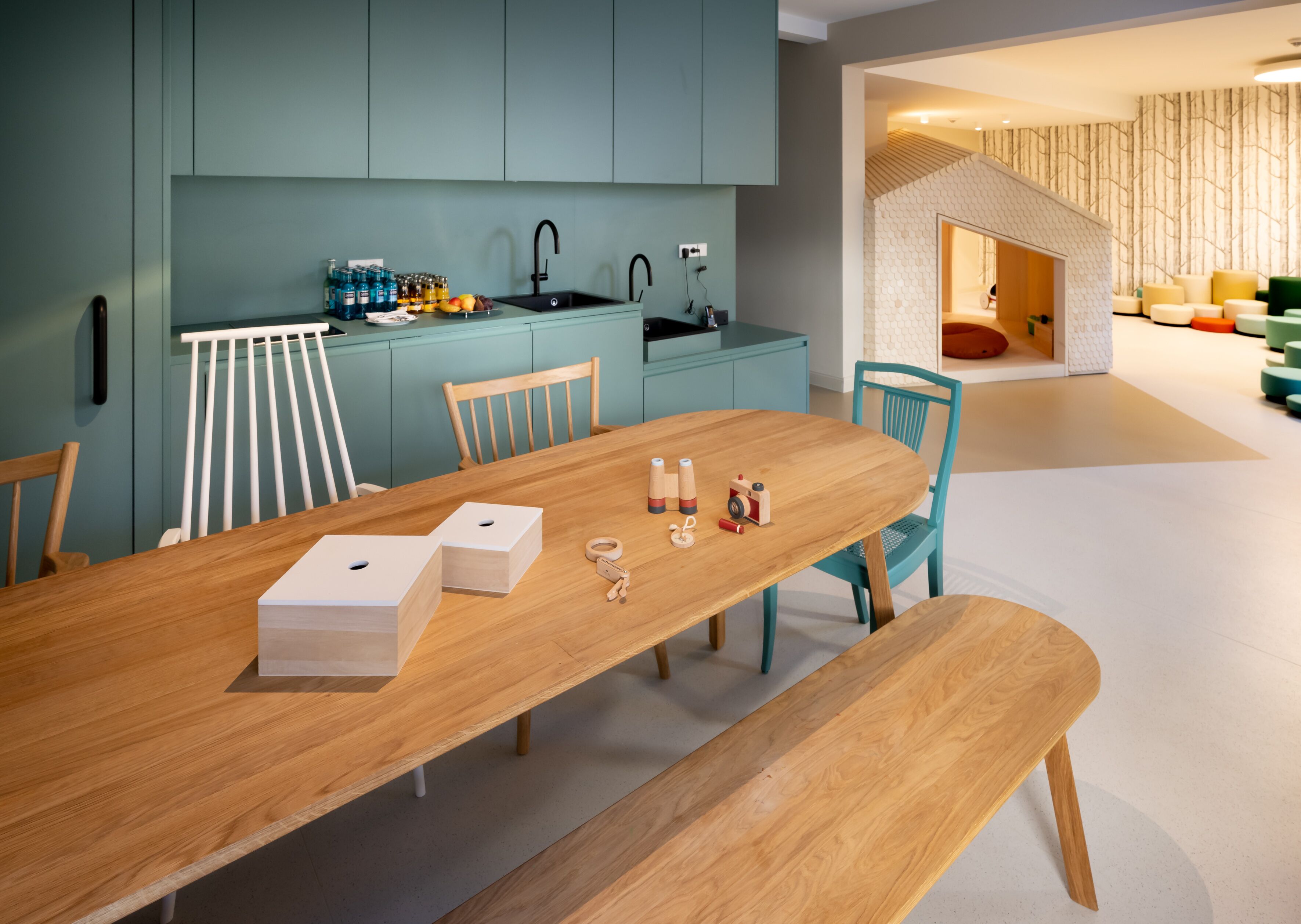 A modern kitchen with teal cabinets, a wooden dining table, and a play area with colorful furniture.