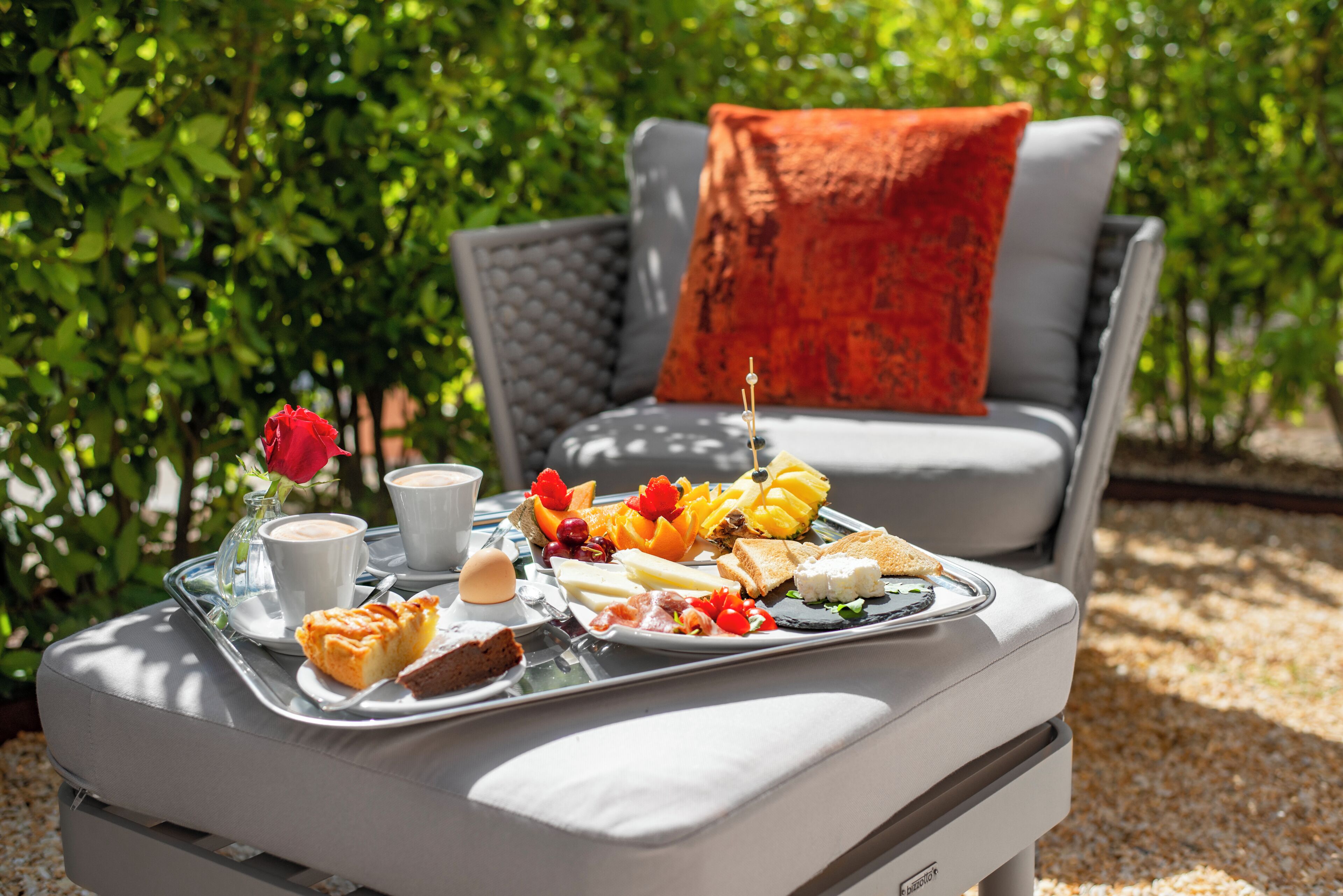 A dining table with a tray of food and drinks, a grey sofa with a red pillow, and a glass vase with a red rose.