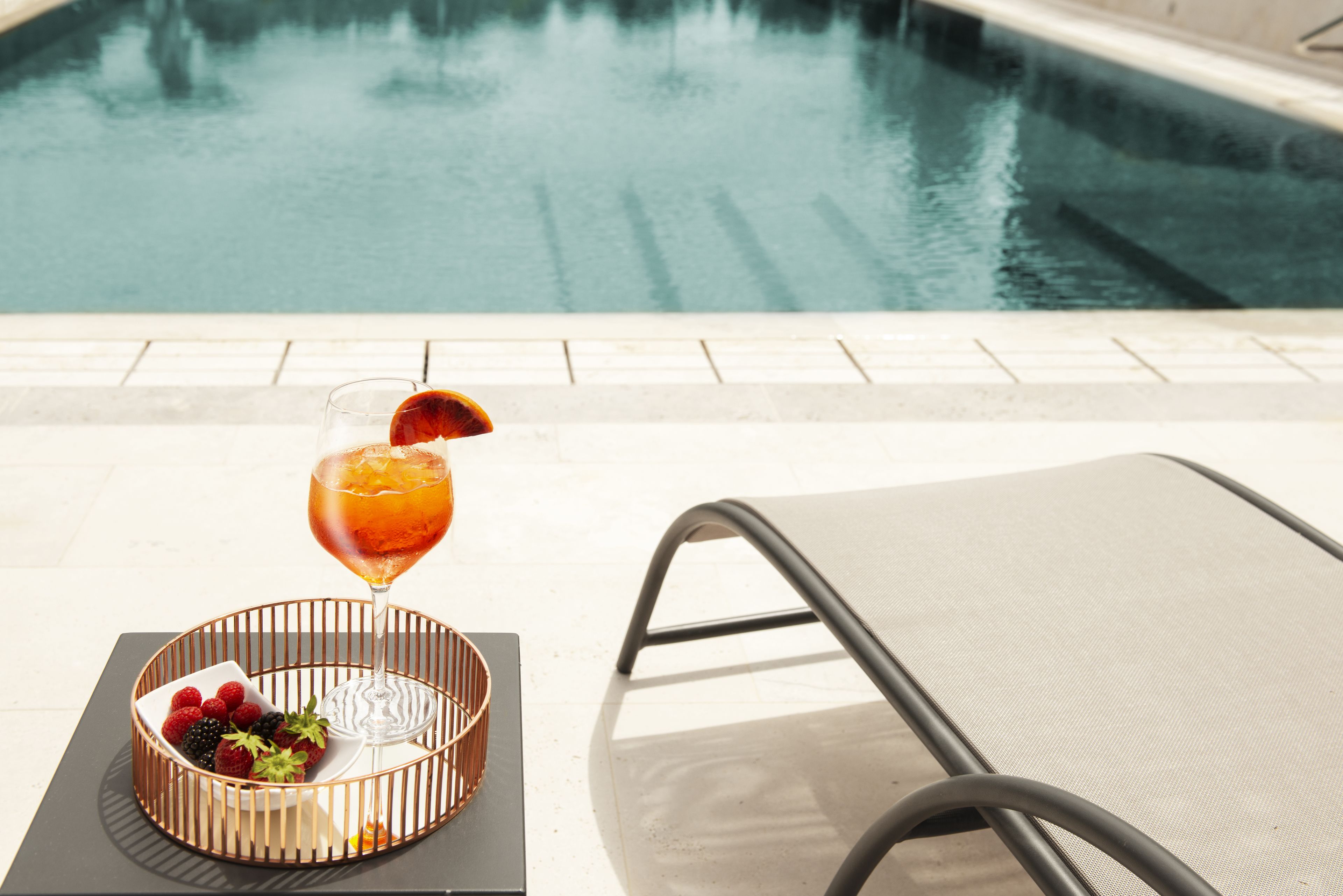 A poolside setting with a lounge chair, a glass of drink with fruit garnish, and a fruit bowl.