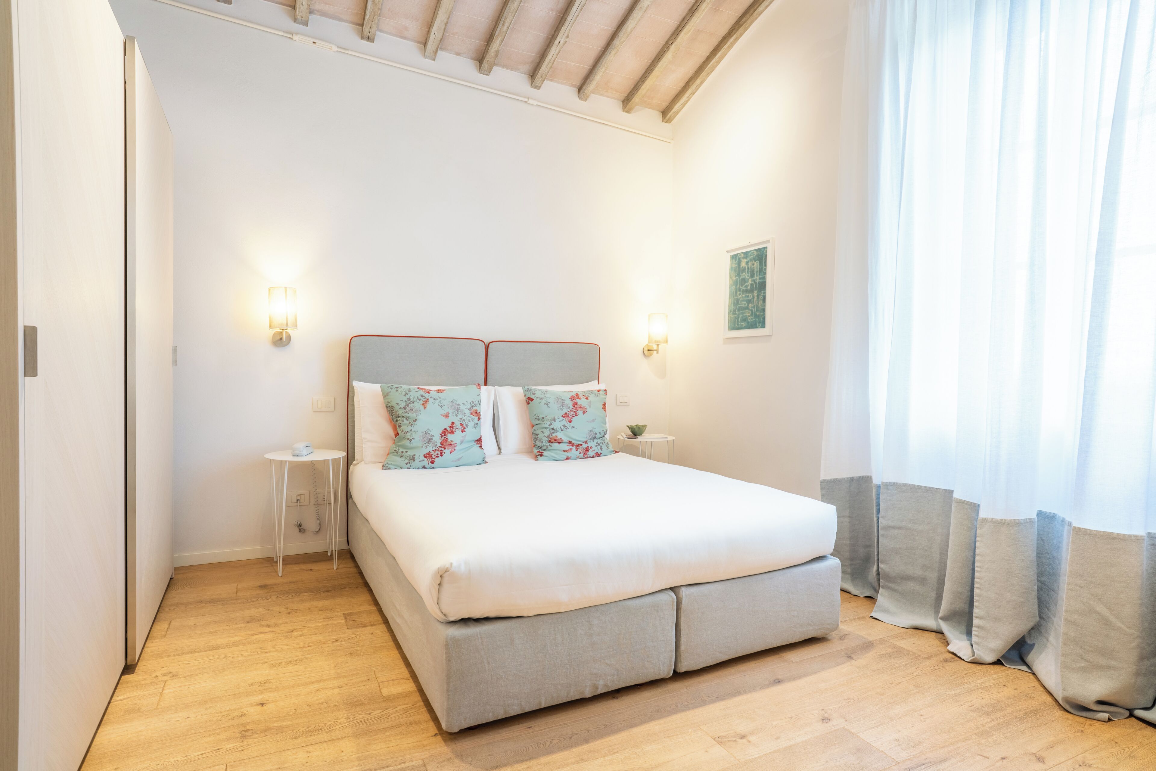 Une chambre à coucher comprenant un lit, des tables de chevet, une commode, une lampe fixée au mur et une fenêtre avec des rideaux.