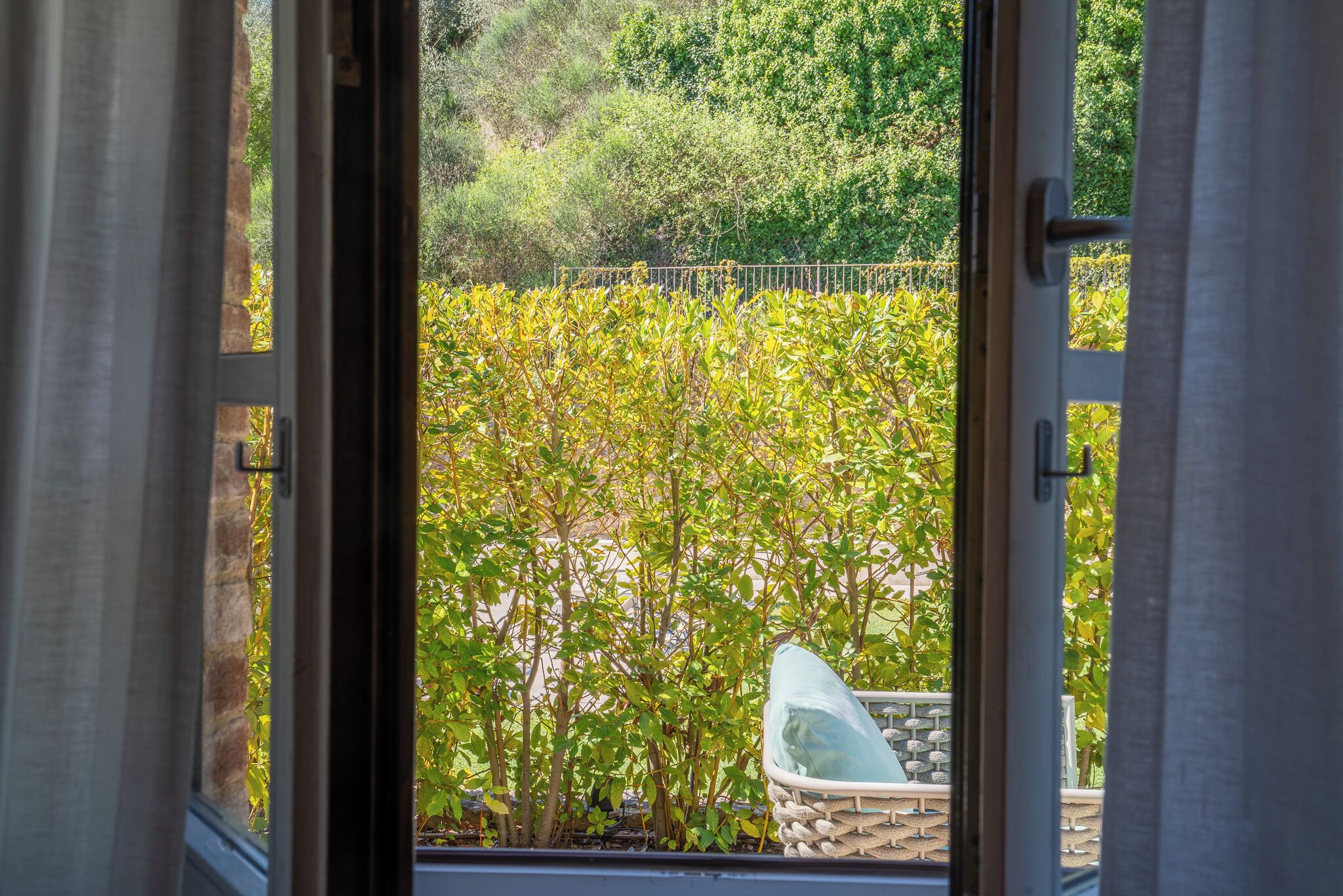 Une vue à travers une fenêtre ornée d’une rambarde en métal et donnant sur un mur de pierre, qui mène à un jardin avec un feuillage verdoyant et une chaise blanche.