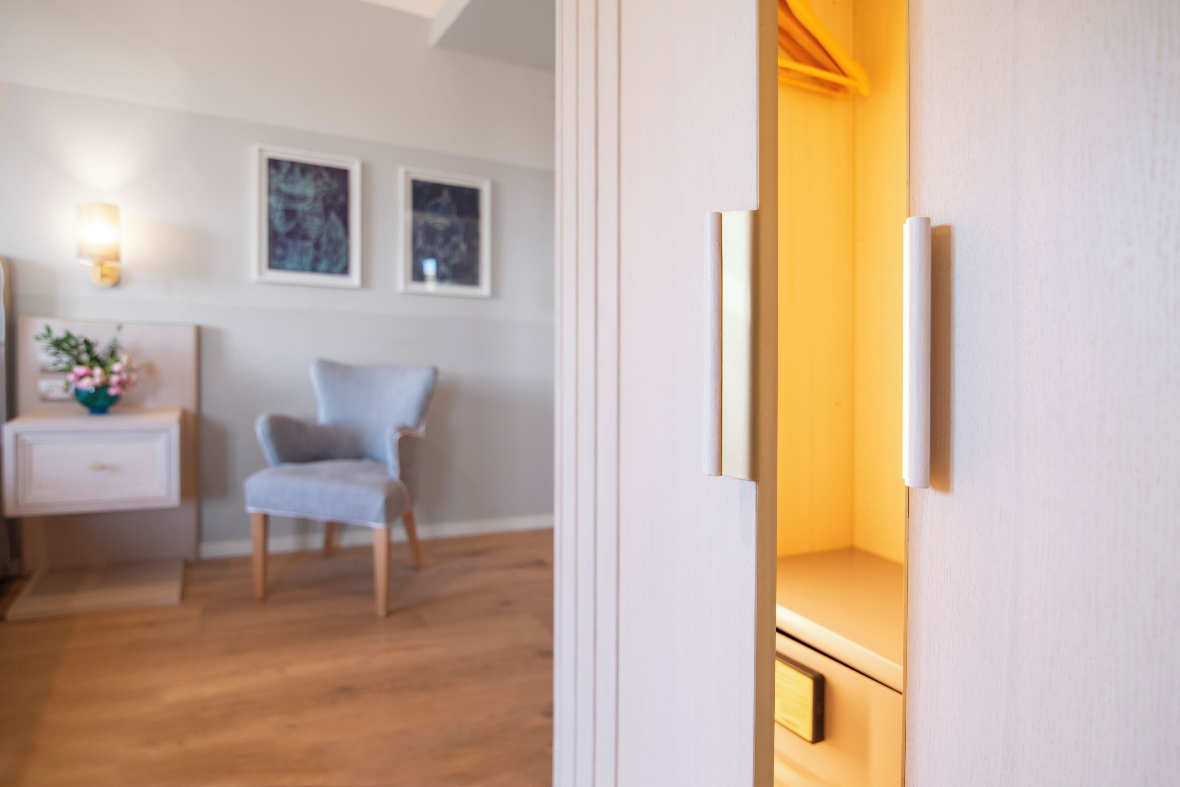 Une pièce avec un plancher en bois, un mur blanc, une chaise, une table de chevet avec une lampe et des tableaux encadrés.