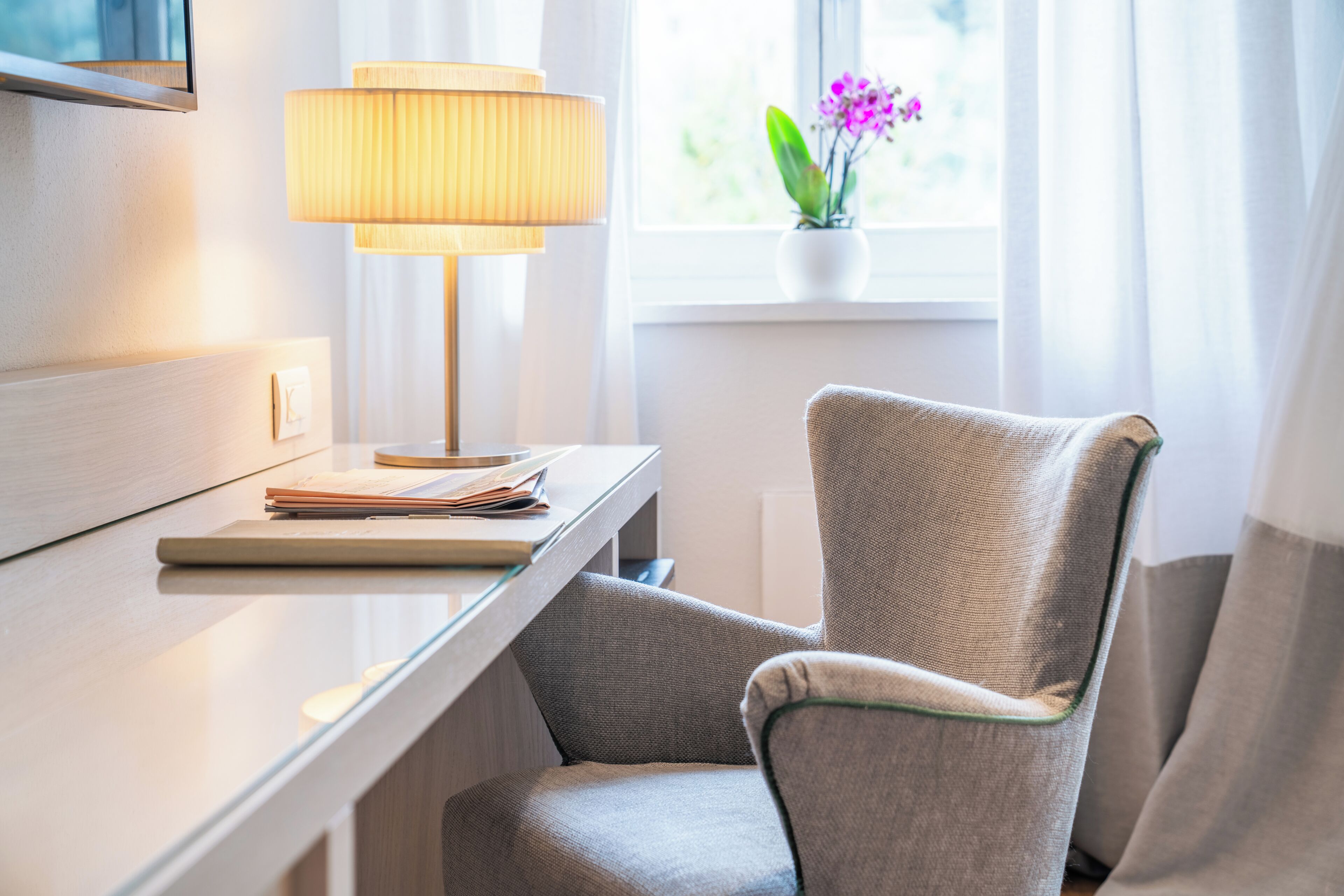 A hotel room with a desk, lamp, chair, and a window with a plant.