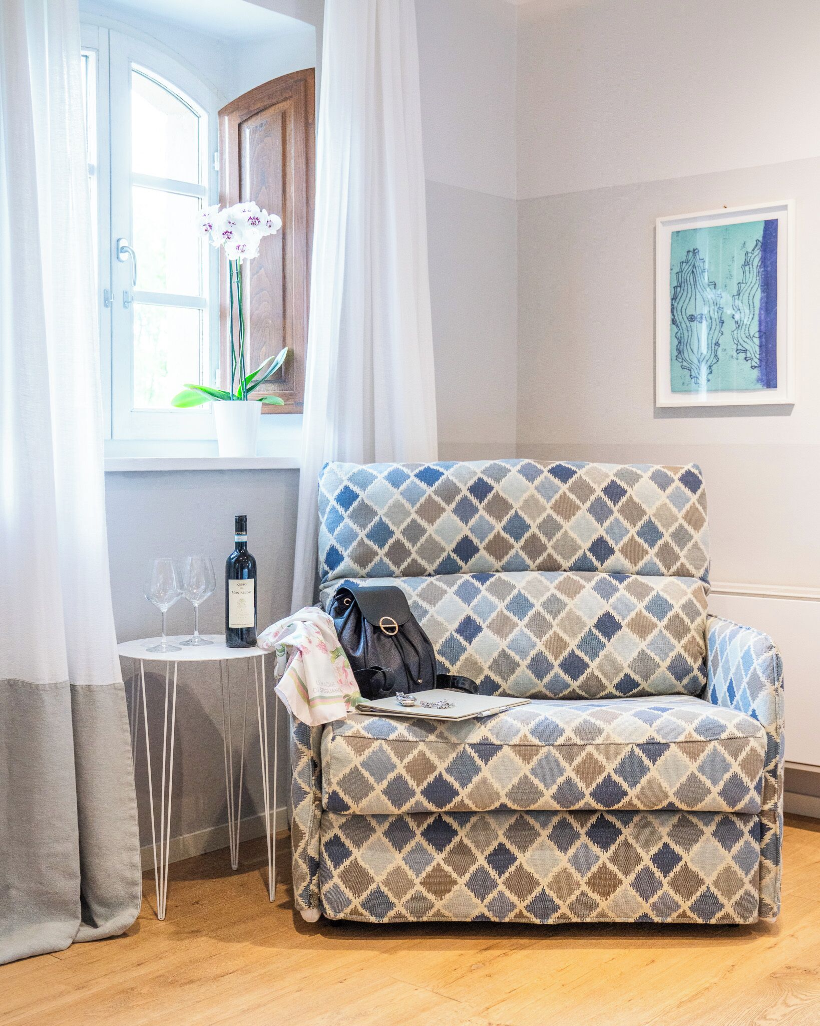 Una habitación acogedora con un sofá estampado, una mesita con copas de vino y una botella, y una ventana con cortinas.
