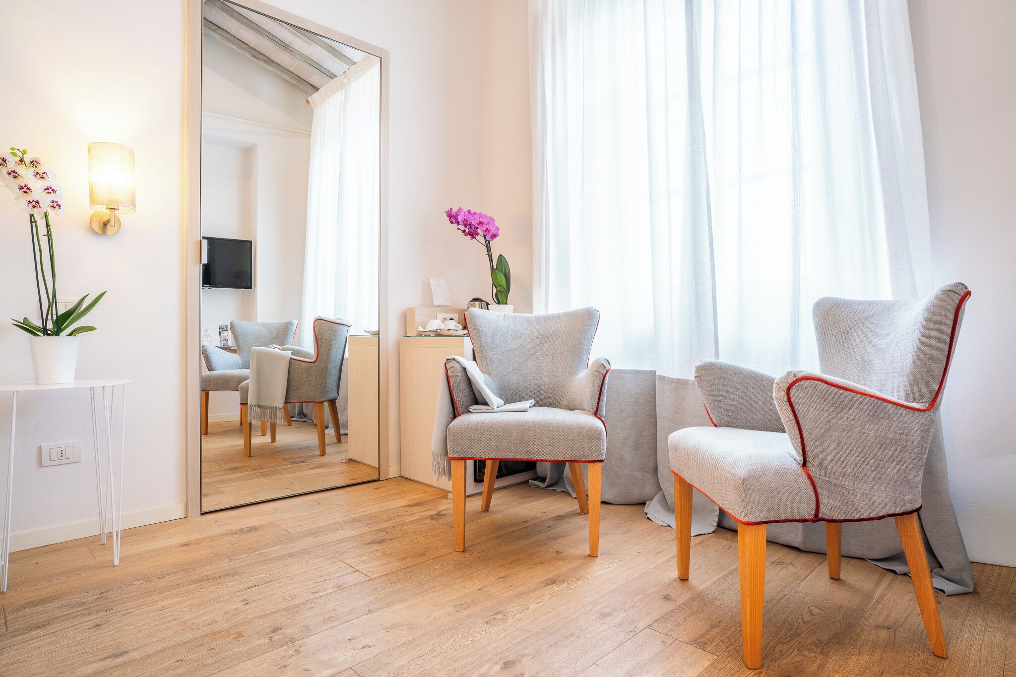Une pièce moderne avec un plancher en bois, deux fauteuils gris à bordure rouge, une console blanche, un miroir et un vase rempli de fleurs.