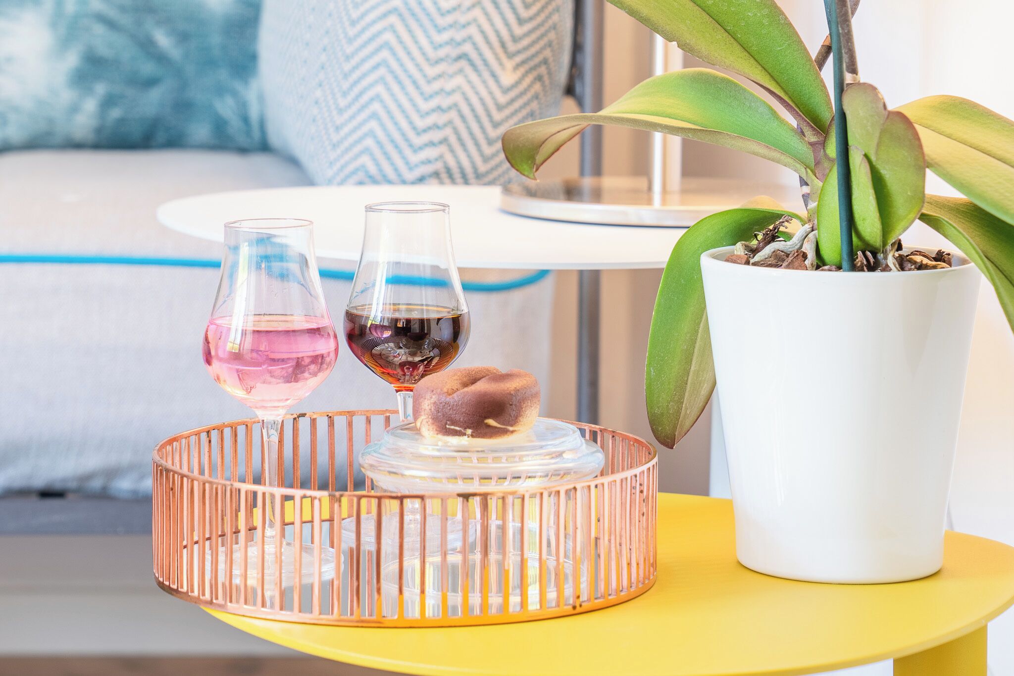Un tavolo giallo con una pianta in vaso bianco, due bicchieri di vino e un vassoio decorativo.