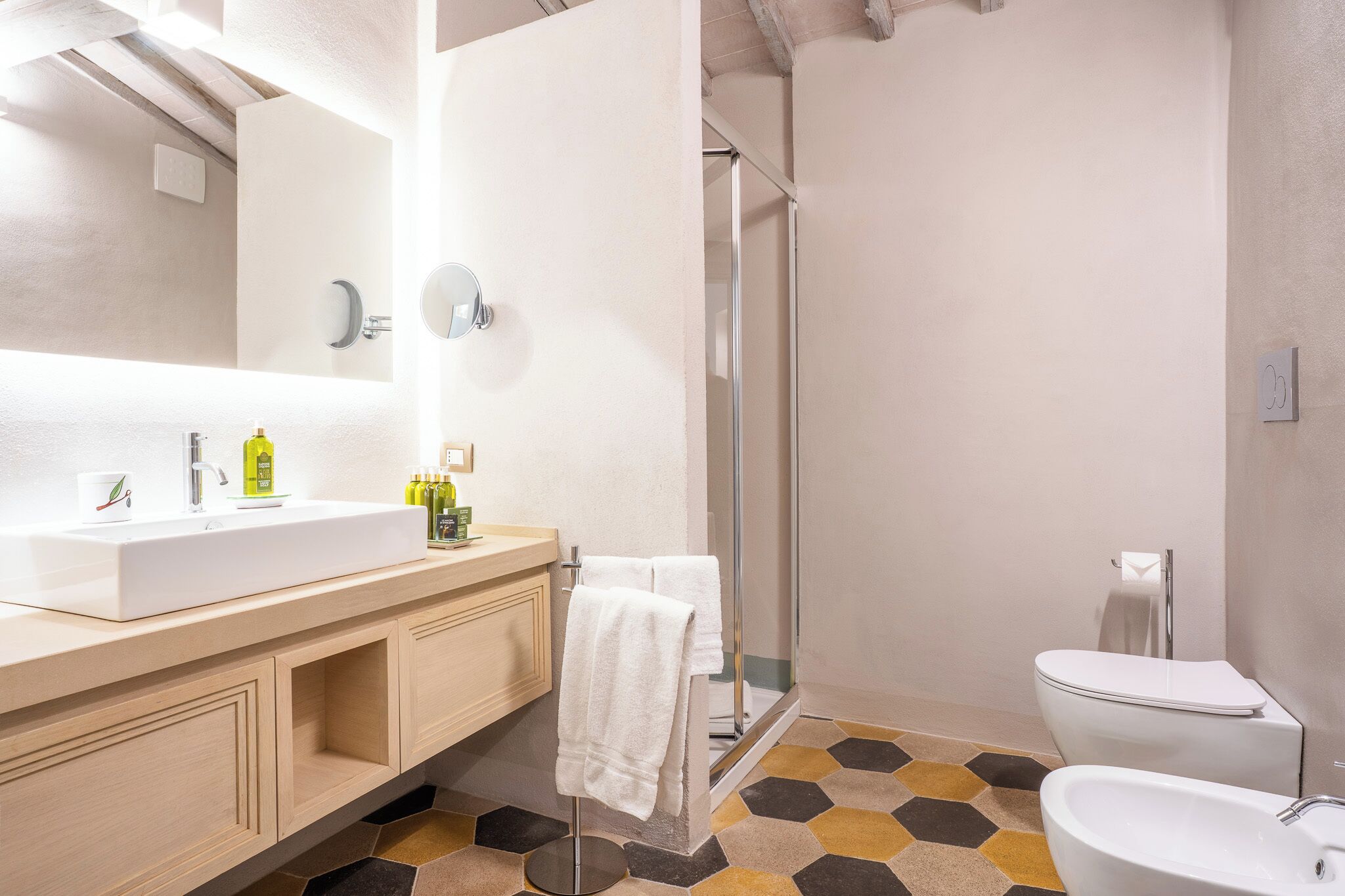 Un baño con lavabo blanco, espejo y inodoro, zona de ducha y suelo con patrón hexagonal.