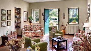 Ein gemütliches Wohnzimmer mit einem blumigen Sofa, einem grünen Sessel und einem Couchtisch. An den Wänden befinden sich gerahmte Bilder und ein Fenster mit Blick auf viel Grün.