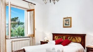 Un dormitorio con un ventanal grande, una cama con almohadillas rojas, un cabecero de madera, una mesita de noche con lámpara y un cuadro enmarcado en la pared.