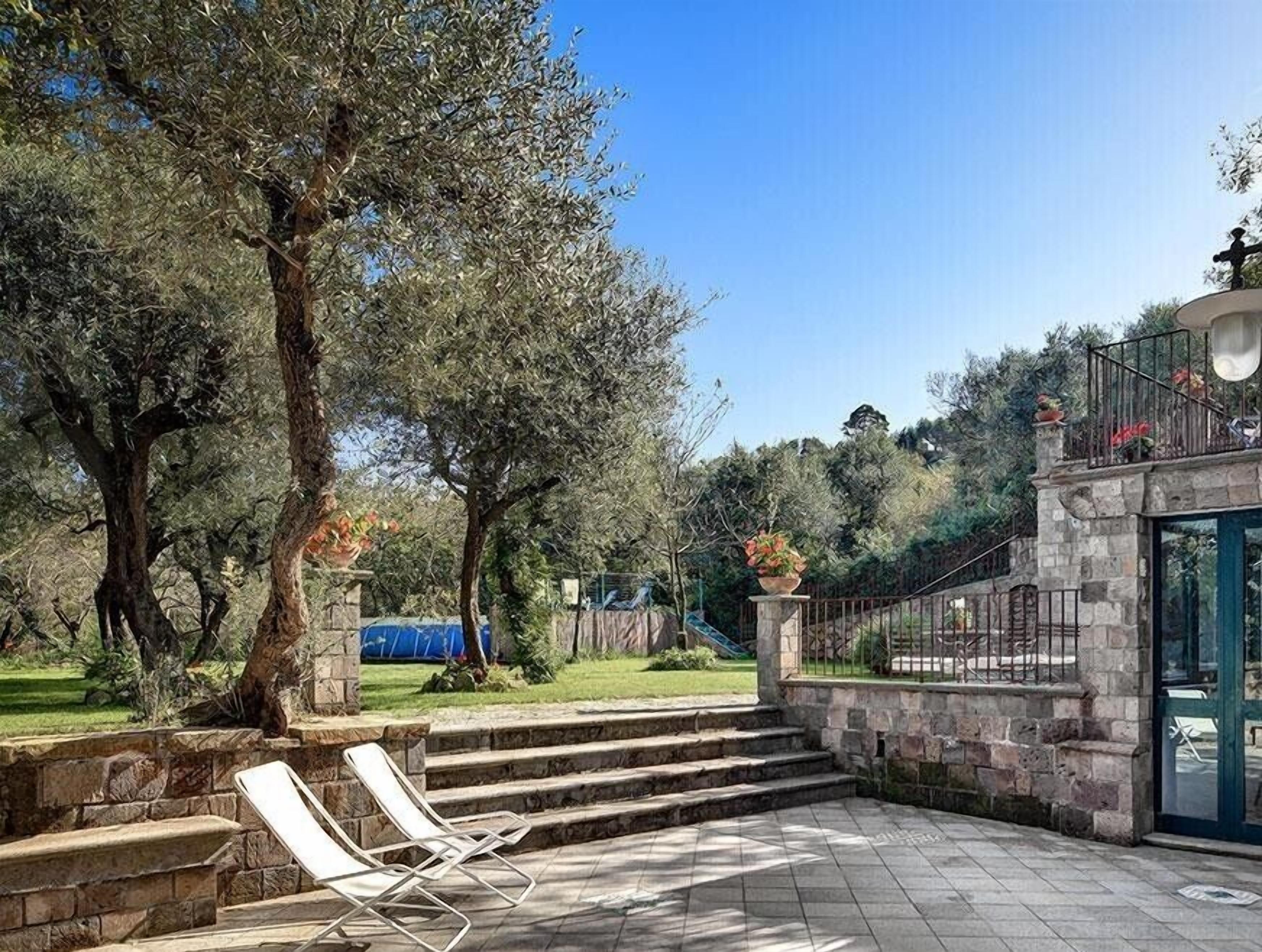 A courtyard with stone steps leading to a covered area, surrounded by olive trees and a stone wall.