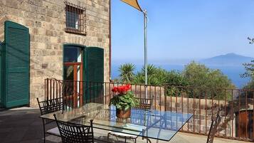 Mesa de vidrio con sillas y una planta en maceta en un balcón con pared de piedra y contraventanas verdes.