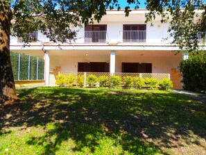 A two-story building with a covered patio area, a fenced garden, and a tree in the foreground.