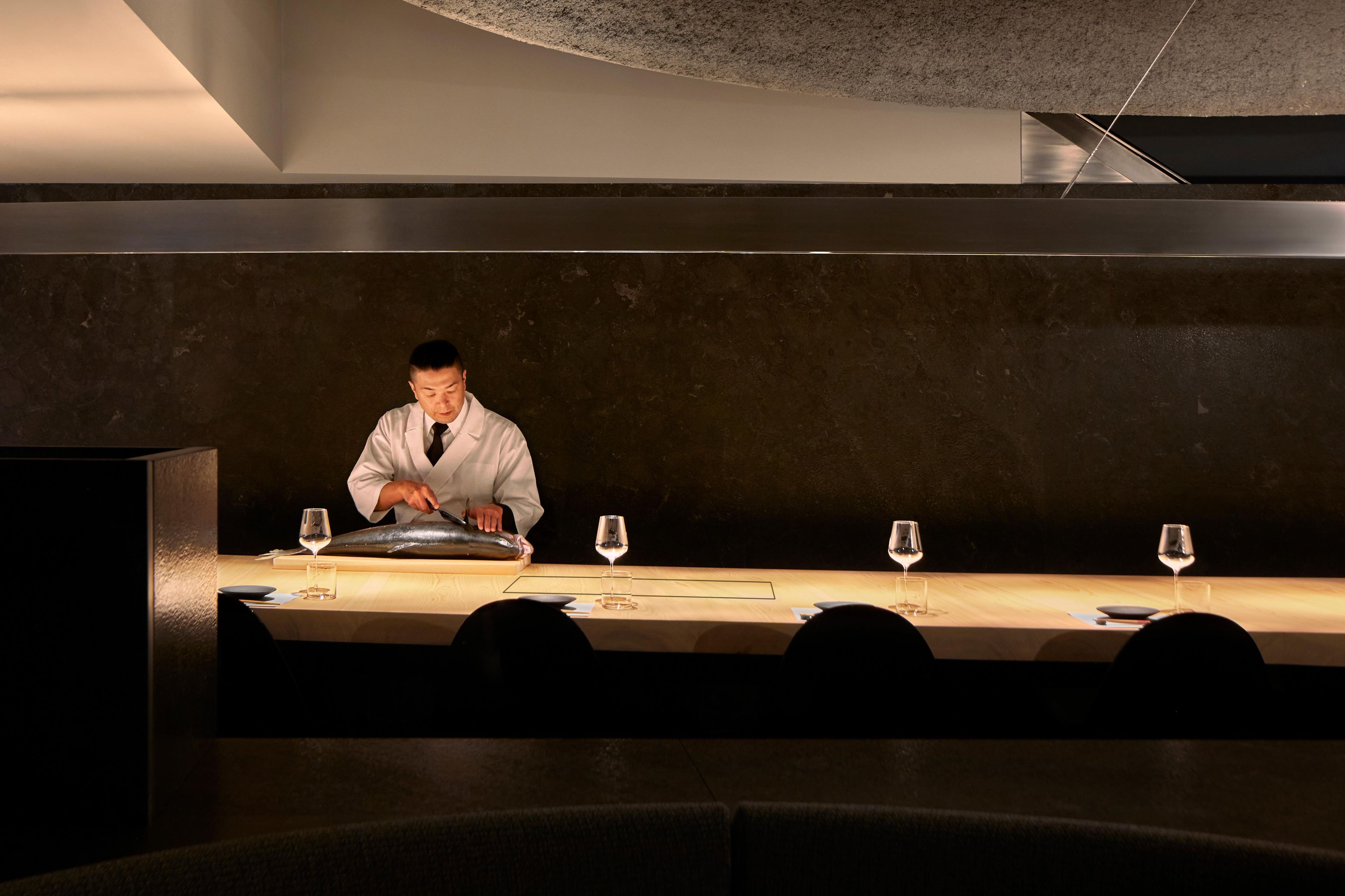 A person in a white coat preparing food at a long table with wine glasses and plates.