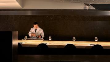 A person in a white coat preparing food at a long table with wine glasses and plates.