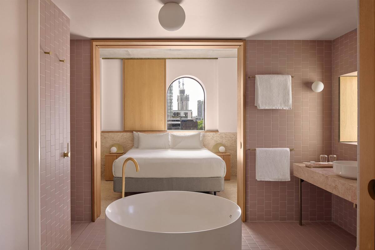 A modern bathroom with a large bathtub, a vanity with a mirror, and a bed with a city view through an arched window.