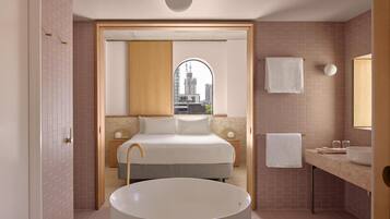 A modern bathroom with a large bathtub, a vanity with a mirror, and a bed with a city view through an arched window.