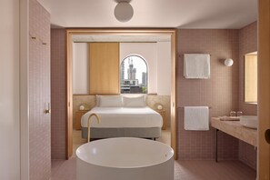 A modern bathroom with a large bathtub, a vanity with a mirror, and a bed with a city view through an arched window.